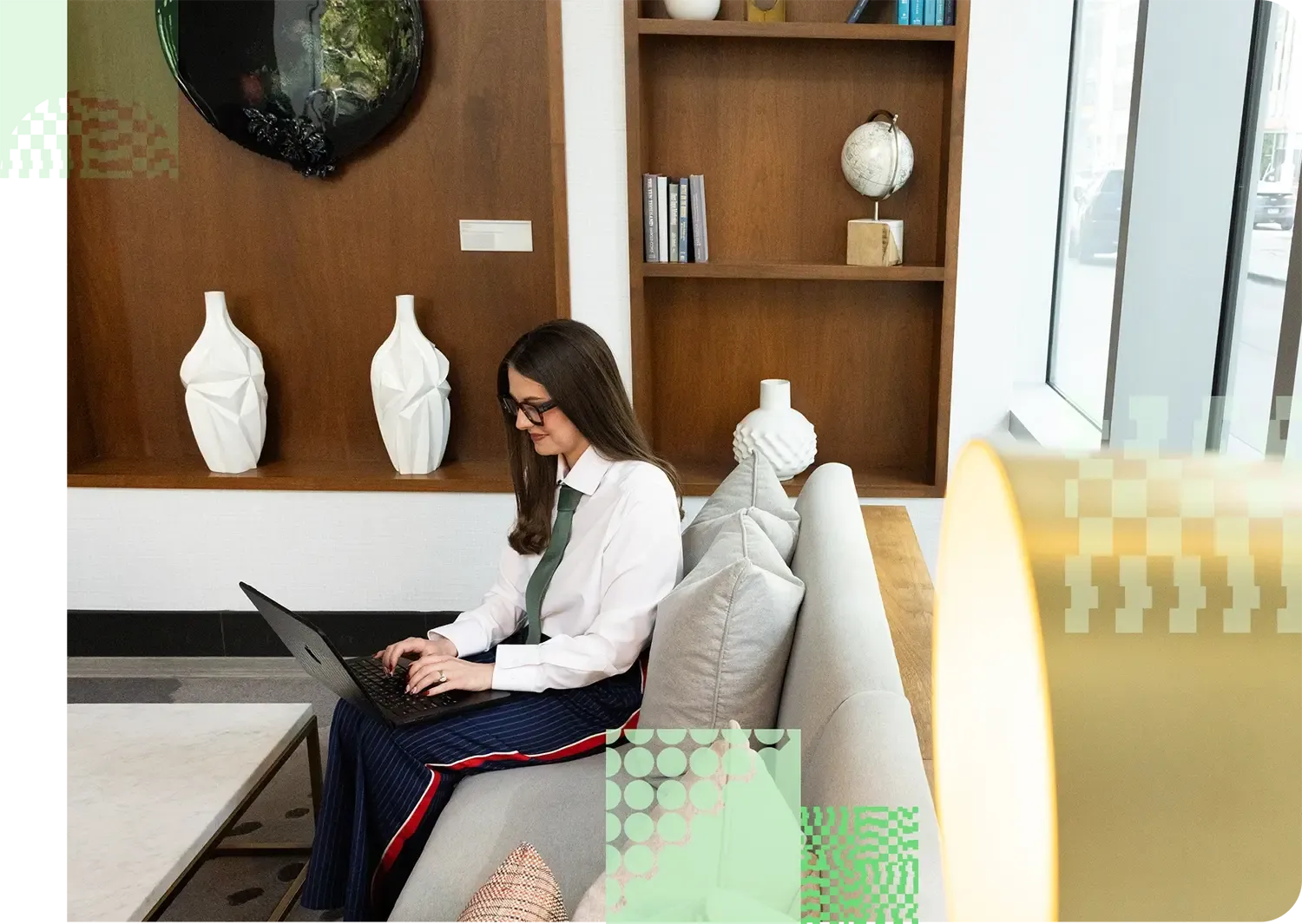 Photo of a woman wearing dark pants, a white shirt with a green tie, and dark glasses sitting on a gray couch in a light-filled room with some dark wood bookcases in the background while working on a laptop on her lap